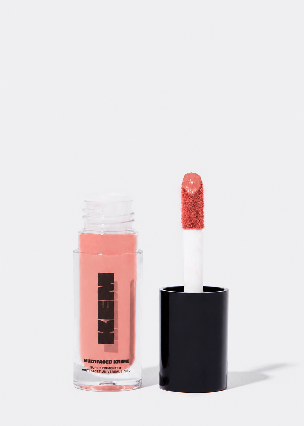A clear bottle of kem matte liquid lipstick with a black cap, shown next to the applicator wand coated in a glittery coral-pink shade, on a clean white background.